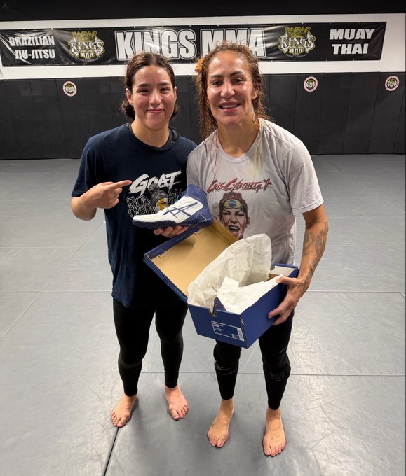 Two women stand on a gym mat in front of a "Kings MMA" banner advertising Brazilian Jiu-Jitsu and Muay Thai, with the younger woman on the left pointing at a pair of athletic shoes while the older woman on the right holds an open blue shoebox. The image illustrates the post's message of gifting training gear to Jaleen Robledo, the rising 22-year-old featherweight talent, as a gesture of encouragement at the end of her training camp.