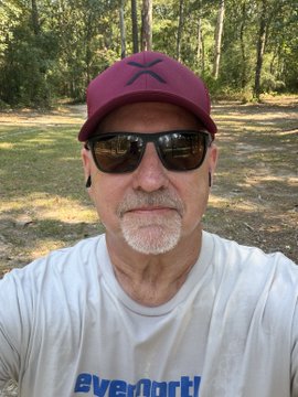A man wearing a maroon cap, sunglasses, and a white "everforth" t-shirt takes a selfie outdoors in a wooded area, his calm expression suggesting a moment of reflection on personal or professional progress.