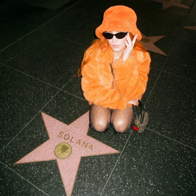 A person wearing an orange fur coat, hat, and sunglasses kneels on the Hollywood Walk of Fame in front of a star engraved with 'SOLANA'.