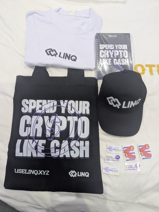 A collection of LINQ-branded merchandise is laid out on a white surface, including a white t-shirt with the "LINQ" logo, a black tote bag featuring the slogan "SPEND YOUR CRYPTO LIKE CASH" alongside "USELINQ.XYZ", a black cap with the LINQ logo, and stickers — likely swag distributed at the Solana Summit Nigeria.