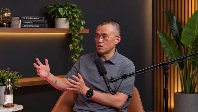 A man with short hair and glasses speaks into a microphone in a studio setting, gesturing with his hands; books titled "THE CHANGING WORLD," "TECHNOLOGY," and "A BRIGHTER TOMORROW" sit on a shelf behind him.