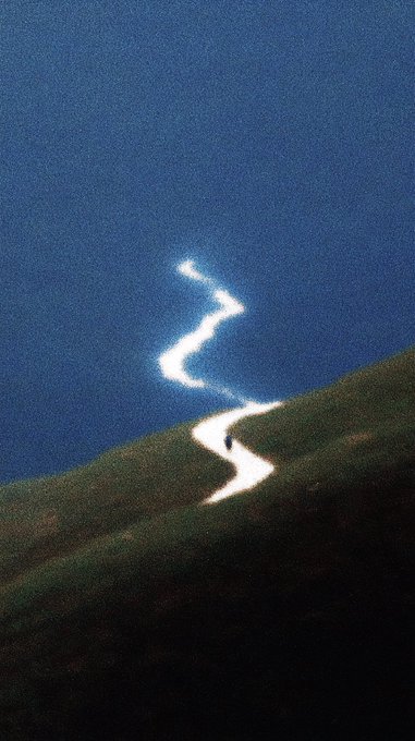 A minimalist, atmospheric digital painting showing a tiny figure walking along a winding white path between a dark green hillside and deep blue sky or water.