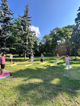 The image shows a group of people practicing yoga outdoors in a park setting. There are at least nine individuals participating in the activity, with some standing and others sitting on mats spread across the grassy area. The scene captures the essence of a Yoga Film Festival, where participants engage in various activities related to yoga, Bitcoin, art, and freedom throughout the day.