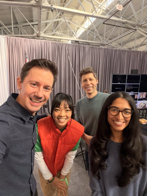Four people pose together smiling in front of a curtained backdrop at what appears to be a tech event, with a monitor visible in the background displaying presentation content. The setting and the accompanying post suggest this was taken at OpenAI's DevDay event in San Francisco, capturing a candid moment among team members or attendees.