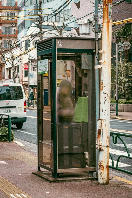 A green NTT phone booth stands on a Japanese street corner next to a rusted utility pole, with a blurred figure inside, a white van, and urban buildings visible in the background.