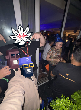 An indoor social gathering shows several people, with one person in the foreground holding up an NFT-style digital artwork of pixelated characters.