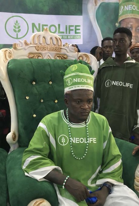 A man wearing a green traditional outfit and cap, seated on an ornate green chair with gold trim, bearing the "NEOLIFE" logo and the phrase "GROW TO THE NEXT LEVEL."