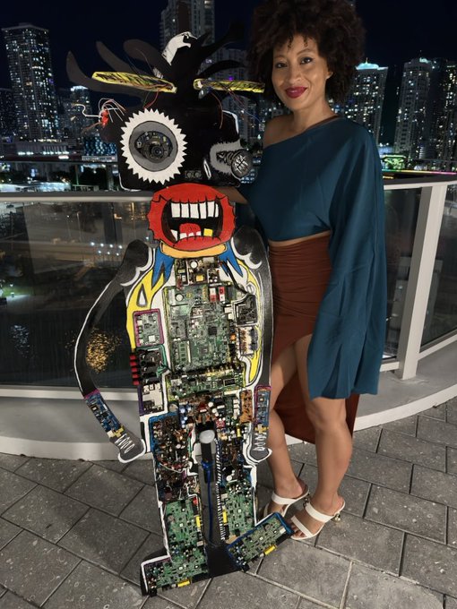 A woman with an afro hairstyle wearing a teal off-shoulder top and brown skirt stands on a balcony at night next to a colorful cardboard cutout character filled with electronic circuit boards.