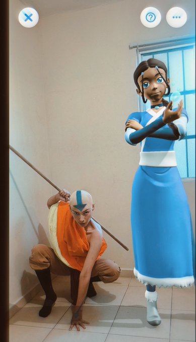 Two people pose in a bathroom wearing Avatar: The Last Airbender costumes, one cosplaying as Aang with a staff and airbender tattoos, and the other as Katara in her blue Water Tribe outfit.