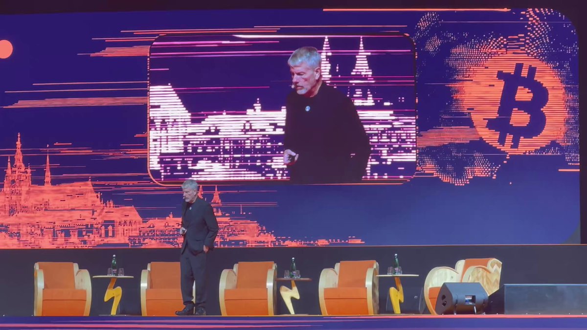 The image features an event where two men are standing behind a stage with a large screen displaying a presentation about Bitcoin. One man appears to be speaking at the event, while the other man is positioned near a chair. The background of the scene includes a cityscape and a building, which adds context to the location of the event. The image also contains text that reads "Saylor at @BTCPrague: Bitcoin is a revolutionary advance. Instead of capital that lasts a hundred years it's a capital that can last a thousand years. Because Bitcoin is deflationary, it has a half-life of infinity." This message seems to be related to the presentation being held on stage and emphasizes the long-lasting nature of Bitcoin as an investment opportunity.