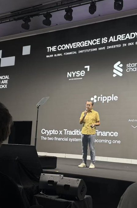 A man in a yellow shirt and jeans speaking on a stage at a conference, with a large screen behind him reading 'THE CONVERGENCE IS ALREADY', along with logos for OKX, NYSE, Standard Chartered, Ripple, and the text 'Crypto x Traditional Finance'.
