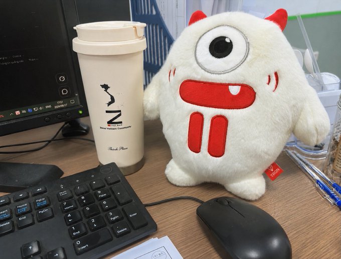 A desk scene featuring a white plush monster toy with one eye and red horns sitting next to an MUJI branded thermos, a computer keyboard, mouse, and monitor.