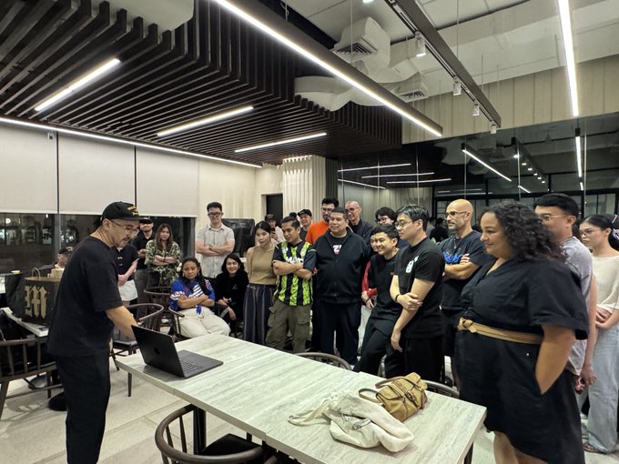 A group of people gathered around a table watching a man present on a laptop in a modern office or coworking space setting.