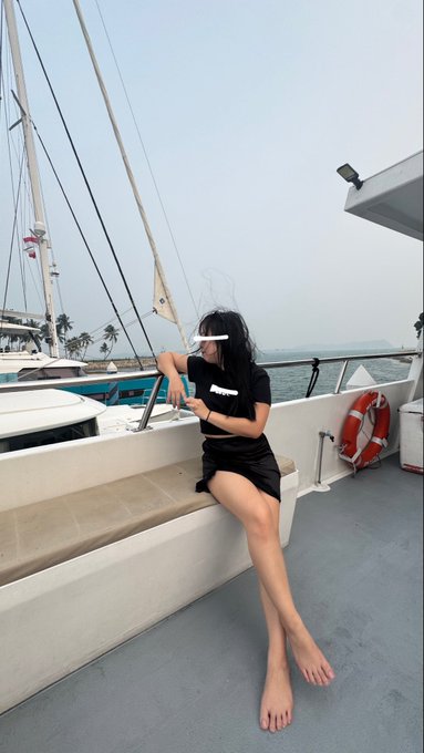A woman in a black top and shorts sitting barefoot on a boat, with her eyes obscured by a white bar, featuring a life preserver and other boats in the background.