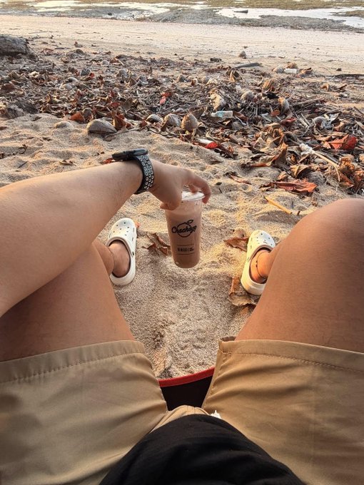 A first-person view of a person sitting on a sandy beach holding a bubble tea drink, wearing tan shorts and Crocs, with debris scattered along the shore.