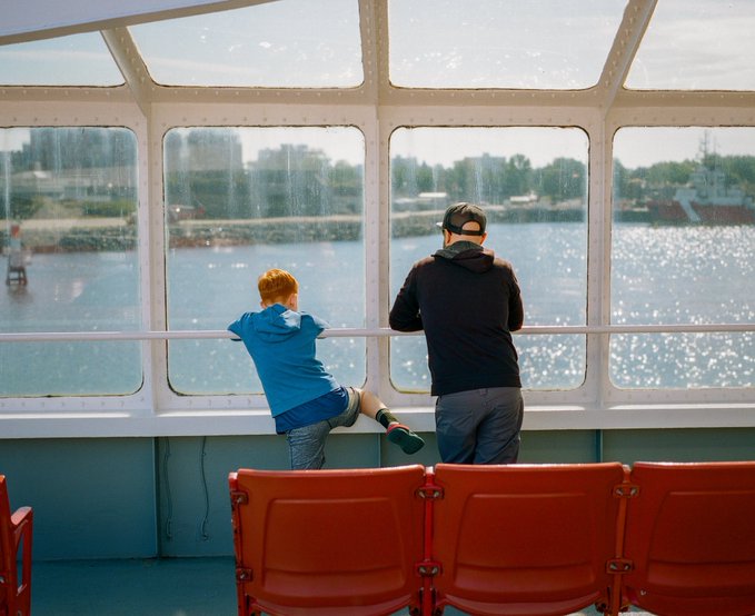 A photograph of a man and a boy leaning on a railing inside a boat with large windows, looking out at a sunlit harbor with a lighthouse and cargo ship visible in the distance, with red seats in the foreground.