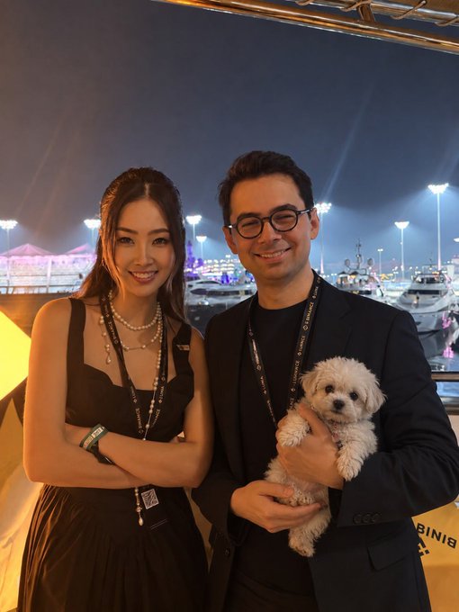 A woman in a black dress with pearl necklaces and a lanyard stands next to a smiling man in glasses and a black blazer who is holding a small white fluffy dog, posing at what appears to be Korea Blockchain Week with boats and event tents visible in the background marina at night.