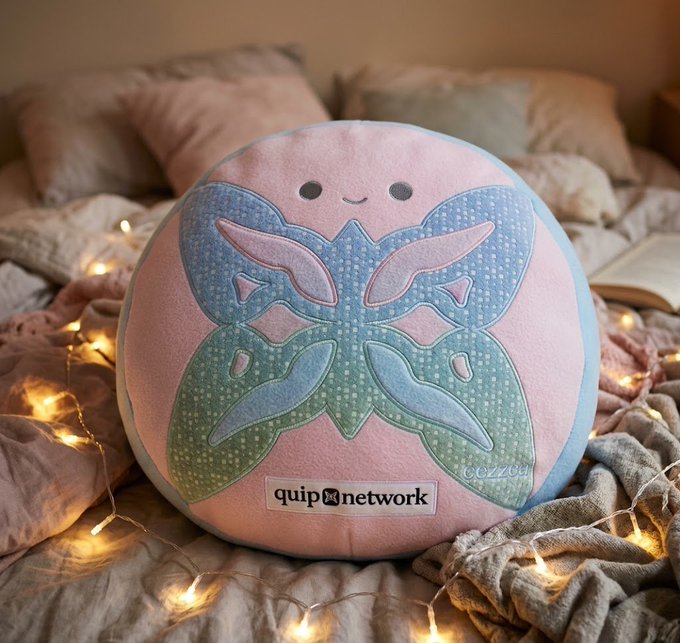 A round plush pillow featuring a smiling face and butterfly-wing design in pastel blue and green hues sits on a bed draped with knit blankets and fairy lights, displaying the "quip network" logo. The image visually represents Quip Network's Quantum Echoes NFT project, which showcases onchain art generated through their QVRF randomness system.