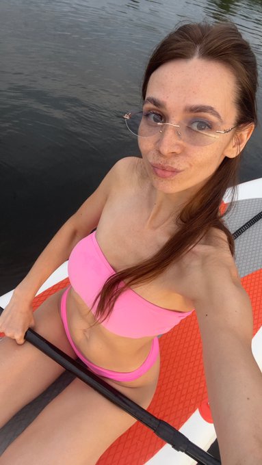 A selfie of a woman in a pink bikini on what appears to be a paddleboard on calm water, wearing tinted glasses.