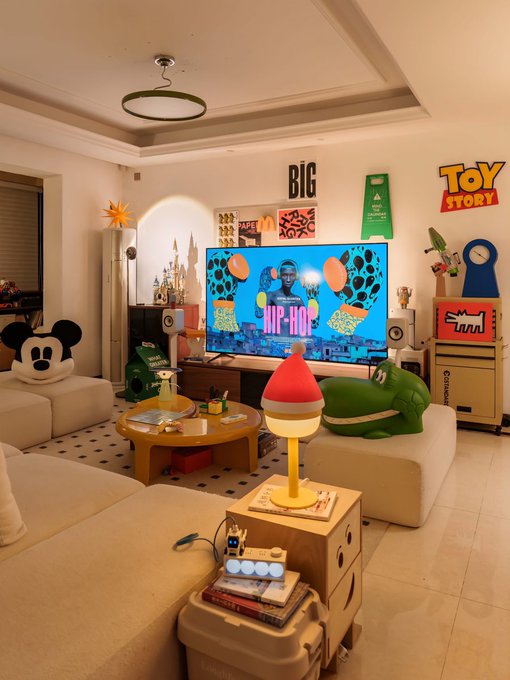 A brightly lit living room decorated with a 'Toy Story' theme, featuring a large TV displaying a hip-hop video, a Mickey Mouse cushion, a green crocodile plush, and various toys and figurines scattered around.