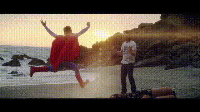 A person in a superhero cape leaps joyfully off a beach rock at sunset while another stands nearby, capturing the moment—visually capturing the ironic "heroic" pose of someone defiantly embracing a losing $WEN stock position.