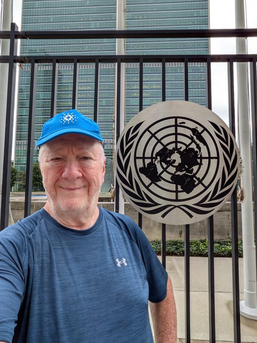 A selfie of a man wearing a blue cap and blue Under Armour shirt, smiling in front of a metal gate displaying the United Nations emblem, with a tall glass building in the background.