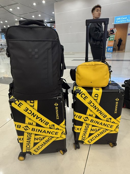 Two suitcases wrapped in yellow tape printed with "BINANCE" and "EXCHANGE" sit in an airport, with a black backpack and yellow bag on top, near a sign reading "SIM Card / Currency Exchange."
