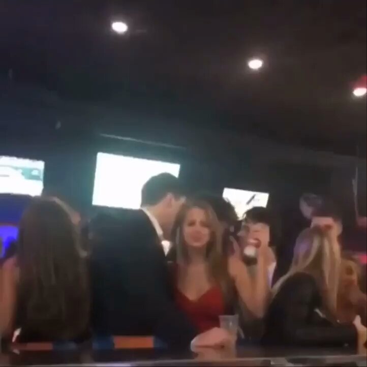 The image shows a group of people gathered around a bar counter, with some sitting at the bar while others stand nearby. A man wearing a suit jacket is leaning over the bar next to a woman who appears to be drinking from a cup. The scene suggests that they are enjoying their time together in what seems to be an informal gathering or party atmosphere.