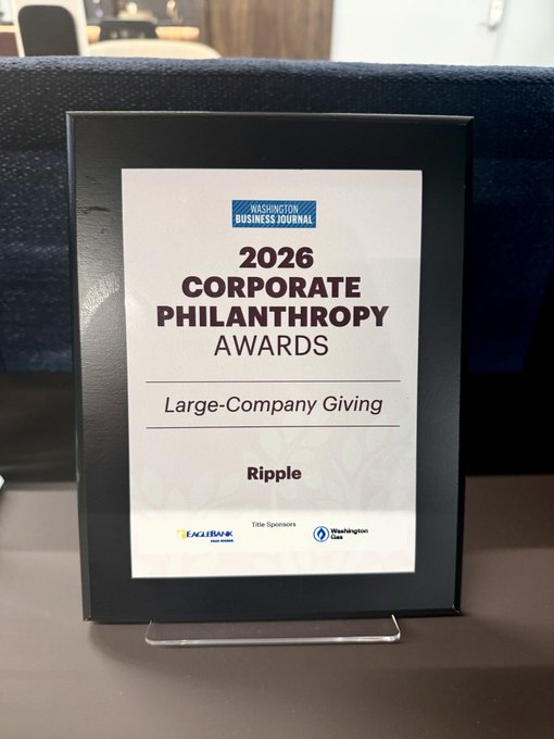 The image features a black plaque with white text that reads "2026 Corporate Philanthropy Awards Large-Company Giving" and "Ripple". The plaque is displayed prominently, possibly at an office or event setting. The background of the image shows a desk area with a chair visible in the foreground.