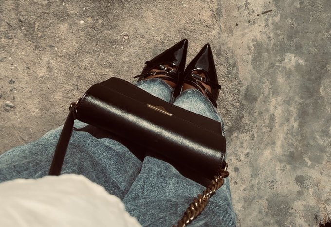 A first-person view looking down at black pointed-toe heels and blue jeans, with a black handbag resting on the lap against a concrete ground.