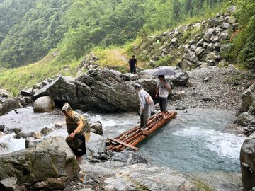 Several people carefully cross a rushing, rocky stream on a makeshift bamboo bridge in a mountainous, forested area, with one person holding an umbrella and another wearing a conical straw hat. The image illustrates the post's caution about the physical demands of outdoor activities like hiking, reinforcing the advice to have accident insurance and sign liability waivers before participating.