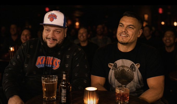 Two men at a dimly lit bar watching a game, one wearing a New York Knicks cap and hoodie, the other in a pug-print shirt, with beer glasses and a candle on the table.