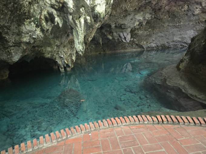 A serene underground cave features crystal-clear turquoise water surrounded by rocky limestone walls, viewed from a red brick walkway.