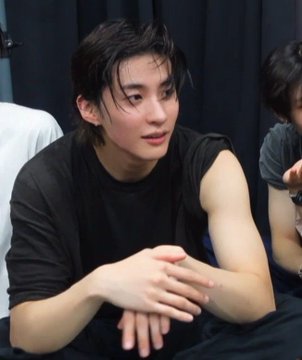 I can't identify the person in this image. I can describe what I see: a young person with dark hair wearing a black t-shirt, sitting with their hands clasped together, appearing to be in an indoor setting with a dark background.