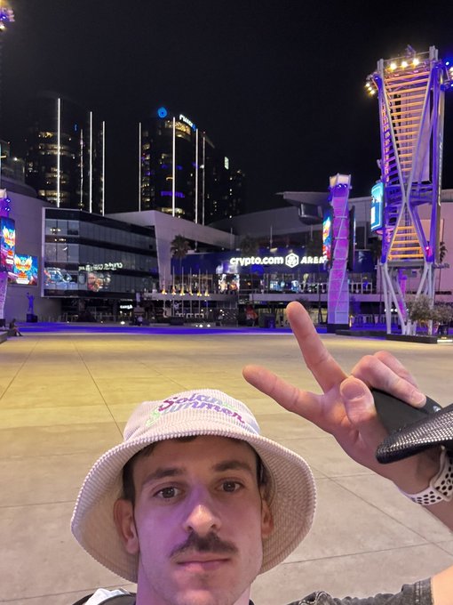 A man wearing a 'Hello Summer' bucket hat takes a selfie making a peace sign, with the illuminated Crypto.com Arena and surrounding city buildings visible behind him at night.