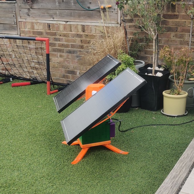 An orange, wooden solar panel stand holding two panels on a green artificial grass lawn, next to potted plants and a brick wall.