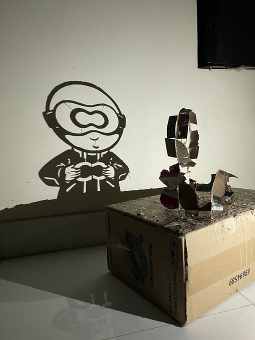 The image features a cardboard box that has been repurposed as a paper mache bird sculpture. The bird is being posed by a person who appears to be wearing goggles, giving an artistic feel to the scene. Surrounding the bird, there are various objects scattered around, including a can of Sprite on its side and another can placed near it.