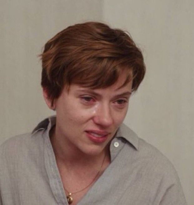A short-haired person with a tearful expression wearing a gray button-up shirt and a gold necklace.