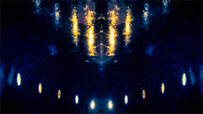 A dark, abstract, mirrored image resembling a symmetrical face or mask made of glowing yellow and orange lights against a deep blue, textured background.