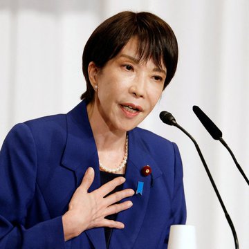 The image features a woman wearing a blue suit jacket and standing behind a podium at an event or conference, likely addressing an audience or participating in a panel discussion. The setting suggests that she could be discussing finance or monetary matters, as there are large sum amounts of money visible on the podium. Nearby, two microphones can be seen, indicating that this is indeed a public speech or presentation. The post also highlights the recent news about Japan's SBI Shinsei Bank allowing customers to convert heir deposit interest into Bitcoin, which could potentially impact the financial landscape and investment strategies in the country.