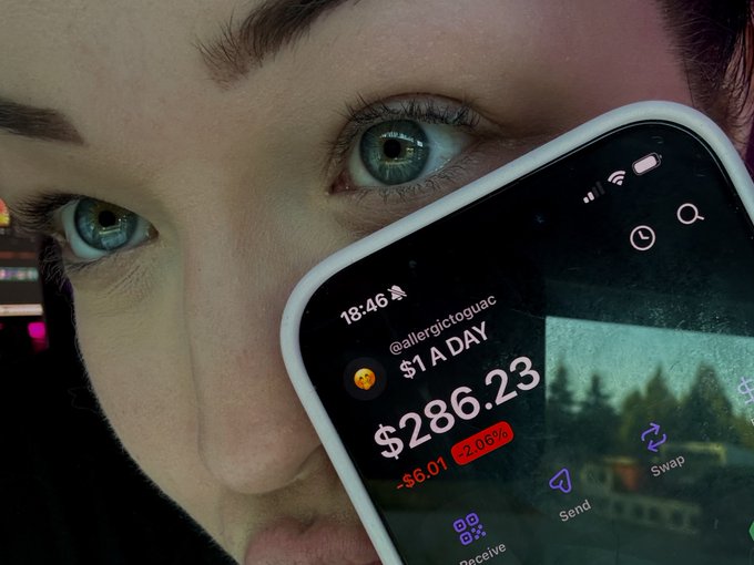 A close-up of a person's face with striking blue eyes partially obscured by a smartphone screen displaying a crypto wallet balance of $286.23, down $6.01 (-2.06%), with the handle @allergictoguac visible.