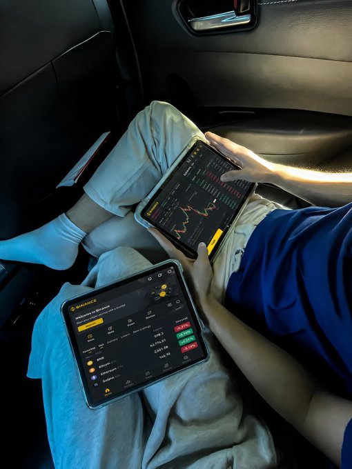 Two people sit in a car using tablets—one displaying the Binance app with cryptocurrency prices (including Bitcoin) and the other showing a stock chart with red and green candles—perfectly capturing how a road trip stays entertaining when Binance is open for live market tracking.