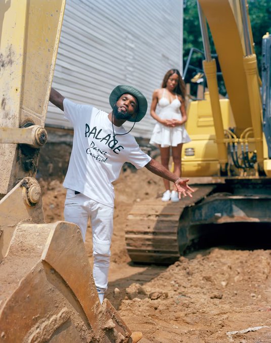 A man wearing a white 'PALACE Dance Central' t-shirt and a bucket hat poses next to a yellow excavator, while a woman in a white dress stands in the background on a construction site.