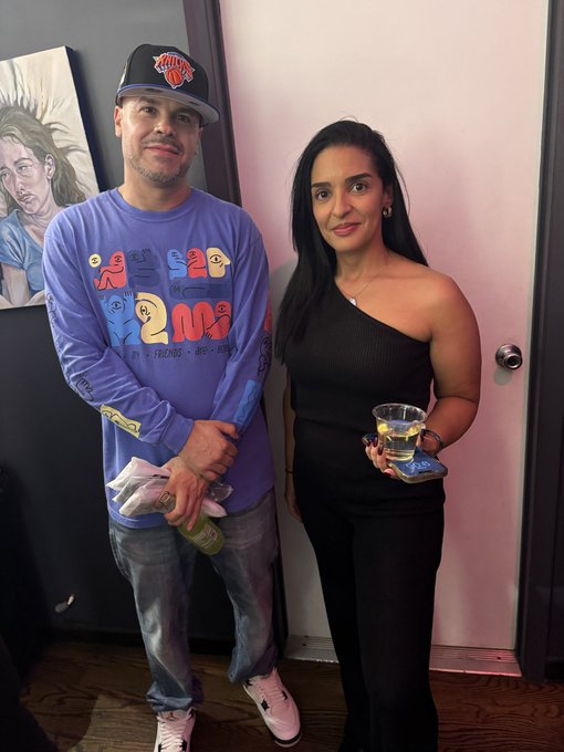 A man wearing a New York Knicks cap and a purple long-sleeve shirt stands next to a woman in a black one-shoulder top holding a drink, posing in front of a painting.