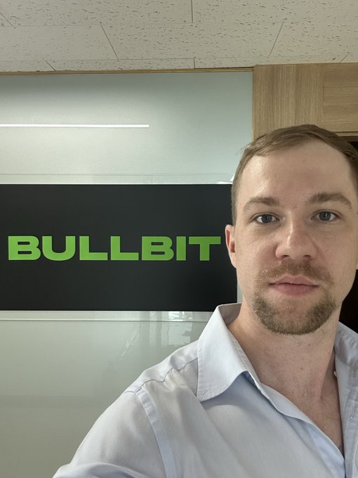 A man in a light blue button-down shirt takes a selfie in an office setting, with a 'BULLBIT' logo sign visible on the glass wall behind him.