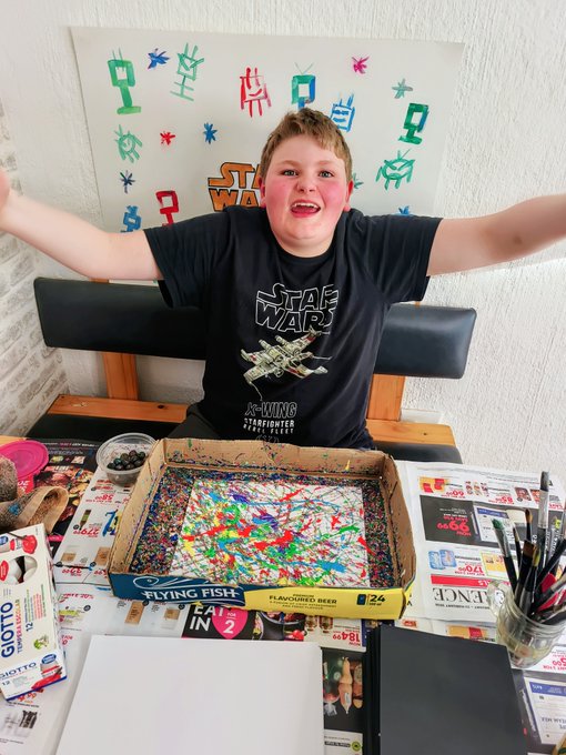 A young artist wearing a Star Wars t-shirt proudly displays his colorful splatter painting in a "Flying Fish Flavorfest Beer 24 Pack" box, surrounded by art supplies. The scene connects to the post celebrating $CAYDEN token support for the budding creator's artistic work.