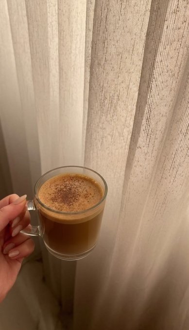A hand holds a glass mug filled with coffee sprinkled with cinnamon, positioned in front of light-colored curtains.