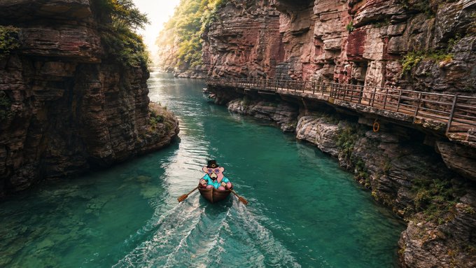Aerial view of a small boat with passengers navigating through a narrow canyon with turquoise water, surrounded by steep rocky cliffs and a wooden walkway built into the rock face.