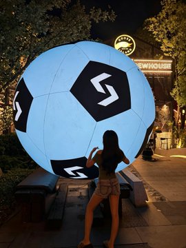 A woman posing at night in front of a large illuminated soccer ball sculpture displaying a 'S' logo, with a 'SHOWORKS' sign visible in the background.