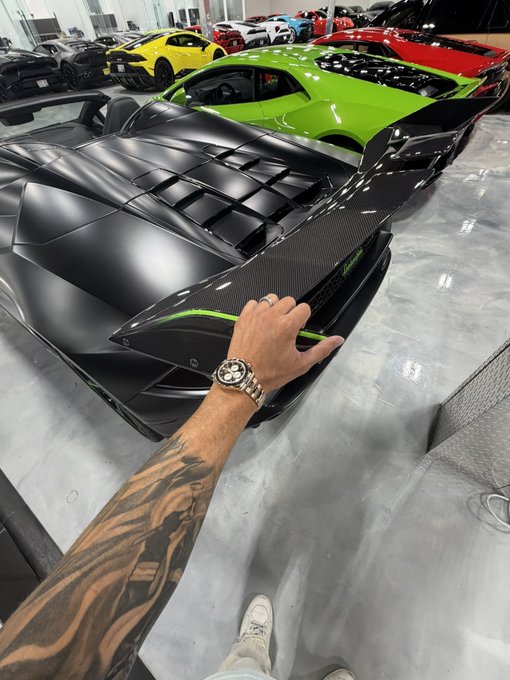 A first-person view of a tattooed arm wearing a luxury watch, with a hand touching the rear of a matte black Lamborghini in a showroom filled with exotic supercars.